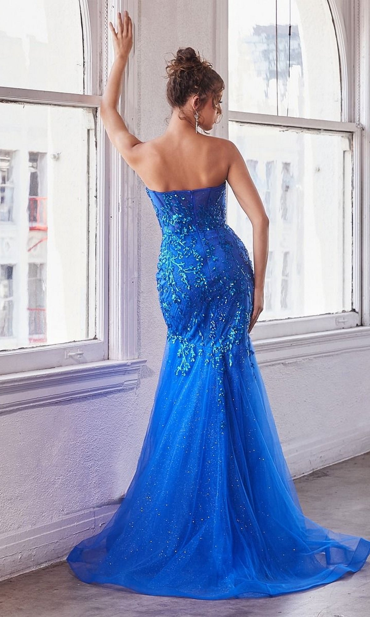 A woman stands by large windows wearing the Strapless Sweetheart Long Mermaid Prom Dress CB139—floor-length, fitted, with sequin embellishments and a tulle train—as she faces away from the camera.